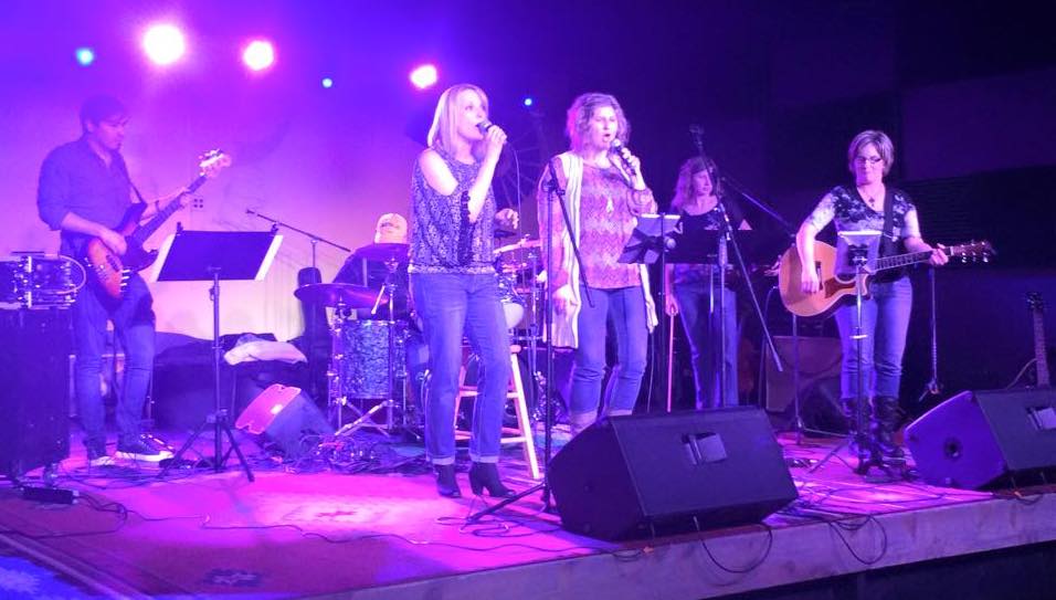 Kelly Crane playing guitar with her band, Chasing Grace doing their thing on a brightly-lit stage.