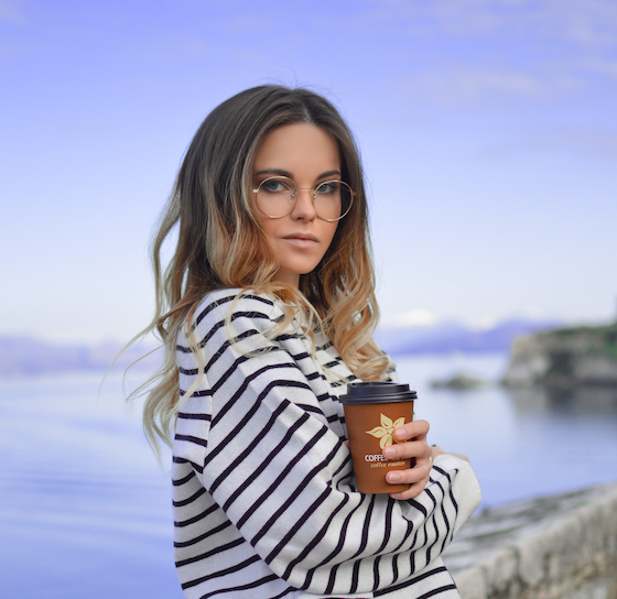 A woman wearing a striped blouse, holding a coffee cup in front of a river.