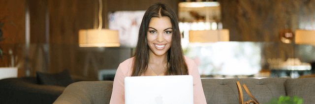 Young woman sitting on a couch, using a computer, smiling at the camera.