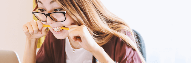 Young woman with brown hair biting down on a pencil, looking at a computer screen.