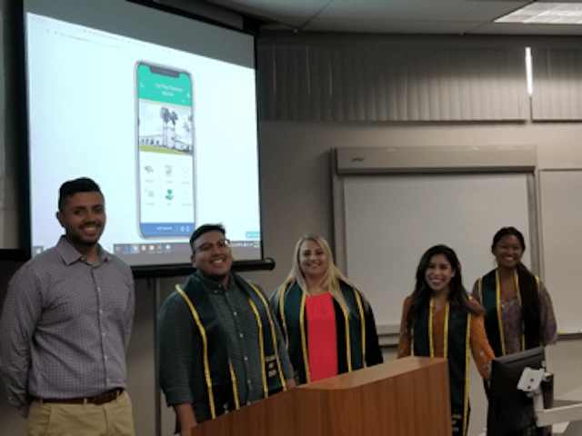 Five students, four of which are wearing graduation stoles, presenting their mobile app design at the front of a room