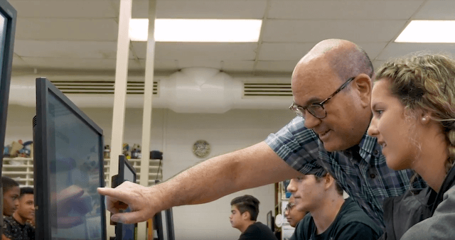 A high school teacher pointing to a student's desktop computer screen in a classroom