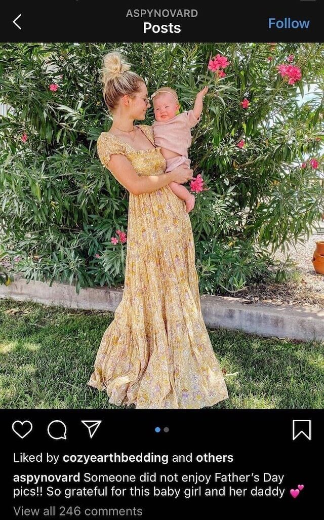 Social media Instagram screenshot of influencer Aspyn Ovard wearing a long yellow dress holding her baby in front of a bush. Aspyn is looking at her baby with a smile and the baby has an arm raised. The caption of the photo says "Someone did not enjoy Father's Day pics!! So grateful for this baby girl and her daddy." The photo has been liked by cozyearthbedding and others.