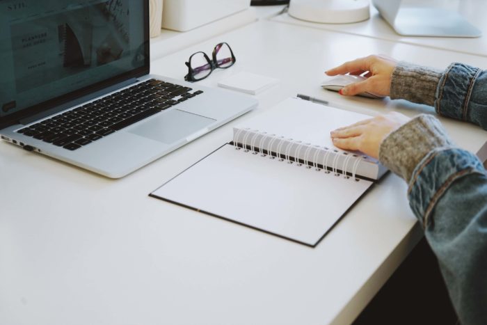 A laptop is open on a table with a blank notebook open in front of it, glasses on the side, and a person has one hand placed on the notebook and one hand placed on a computer mouse.