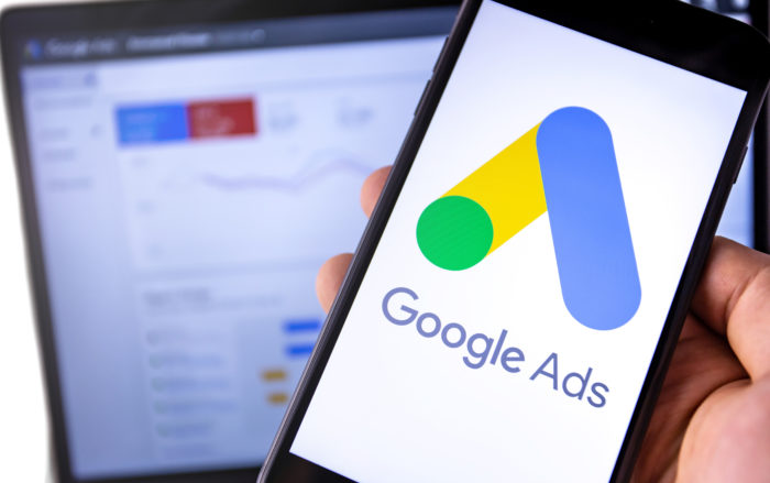 Hand holding cellphone with Google Ads logo on the screen. Computer in background.