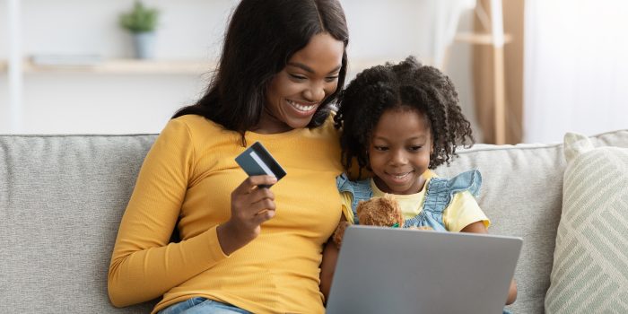 Happy Black Mother And Little Daughter Shopping Online With Laptop Computer And Credit Card While Relaxing On Couch At Home, African American Mom And Female Child Enjoying Purchasing From Internet